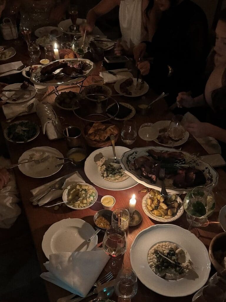 A candlelit table mid-feast at Gigi's in Paris, covered in sharing platters of slow-roasted meat with rosemary, risotto, roasted potatoes, greens, flatbreads and sauces on mismatched vintage-style plates, with wine glasses, candles and hands reaching in around the table.