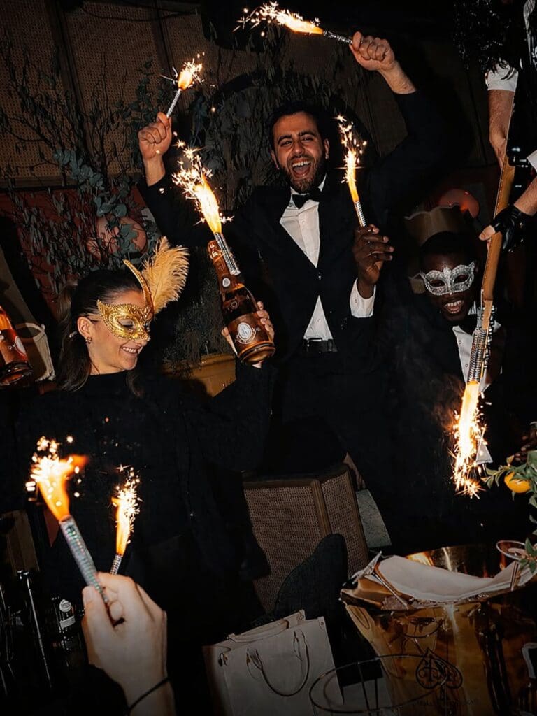 Staff in black tie and masquerade masks holding sparklers and champagne bottles during a late-night celebration at Mamamia in Paris, with arms raised, a gold champagne bucket in the foreground and trailing greenery in the background.