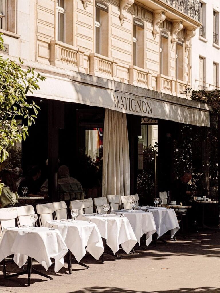 The sun-dappled terrace at Matignon in Paris, with white tablecloth-covered tables set with wine glasses and cutlery beneath a branded awning, dappled shade from the trees, linen curtains at the entrance and an ornate Haussmann-style building facade above.