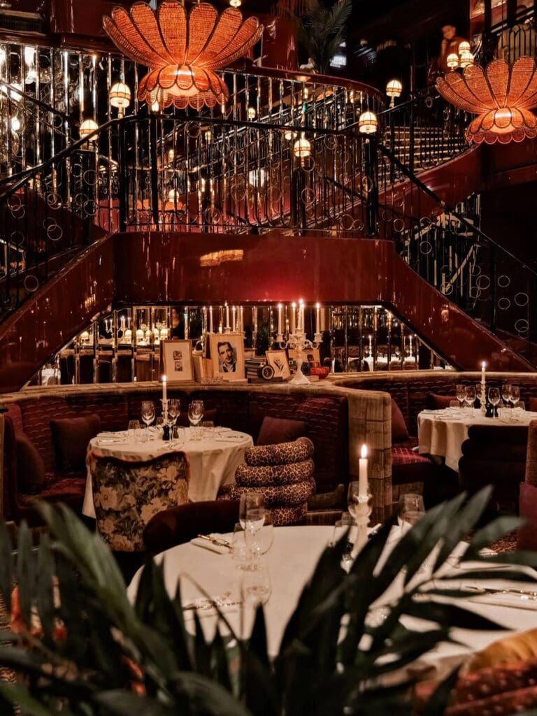 The lavish, candlelit interior of Mondaine de Paris, with white tablecloth-covered tables set with wine glasses and tall candelabras, deep burgundy velvet booth seating with leopard print and floral cushions, framed vintage photographs on the shelves, a sweeping wrought iron staircase with glossy red lacquered panels, oversized orange scalloped pendant lights, mirrored walls and tropical plants in the foreground