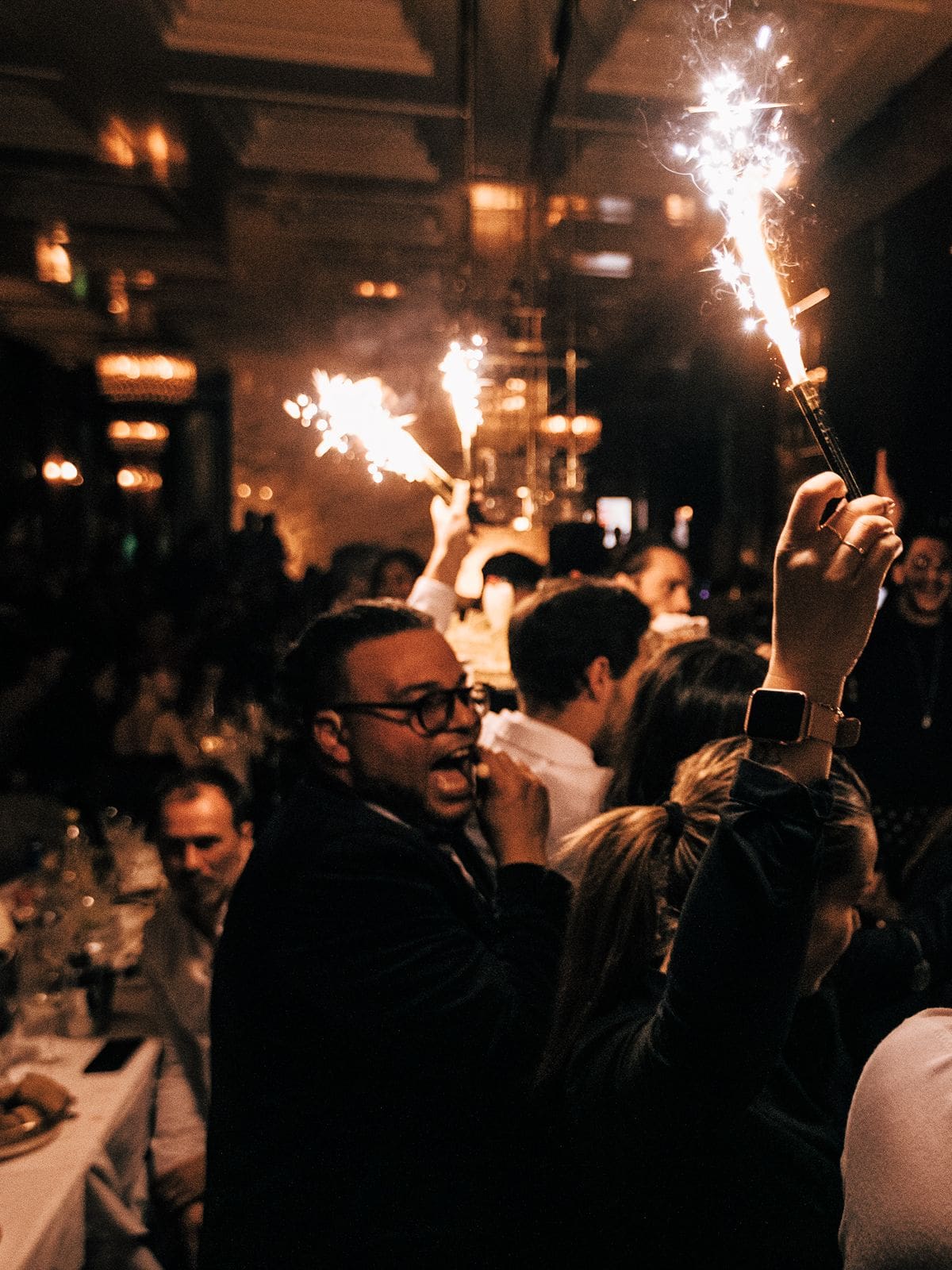 Diners holding sparklers in the air during a late-night celebration at NOTO in Paris, with a man in glasses and a dark blazer cheering in the foreground, a packed dining room behind him, and warm amber lighting and ornate fixtures glowing through the smoke.