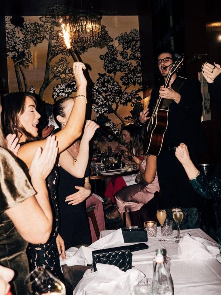 Women dancing and holding sparklers at their table at NOTO in Paris while a guitarist performs beside them, with champagne flutes on the white tablecloth, a dramatic gold and green chinoiserie mural on the wall behind, and a crystal chandelier overhead.
