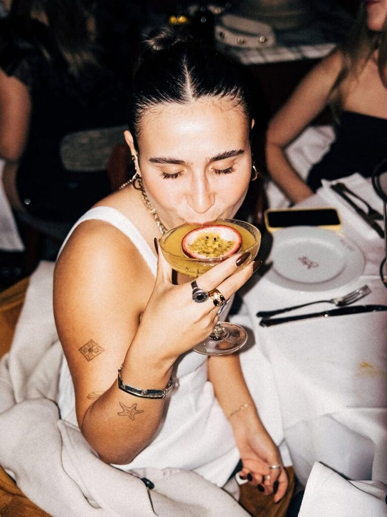A woman in a white vest sipping a passionfruit martini from a coupe glass at NOTO in Paris, with a flash-lit, end-of-the-night feel, white tablecloths and other diners visible in the background.