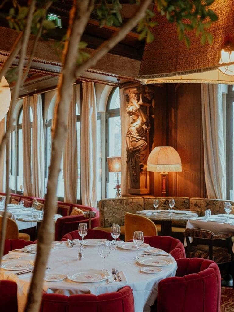 The opulent dining room at Victoria in Paris, with white tablecloth-covered round tables set with wine glasses and branded plates, deep red scalloped velvet chairs, a large classical sculpture of a woman holding grapes, floral tapestry banquette seating, fringed pendant lampshades, flowing linen curtains at tall arched windows, and indoor trees framing the foreground.