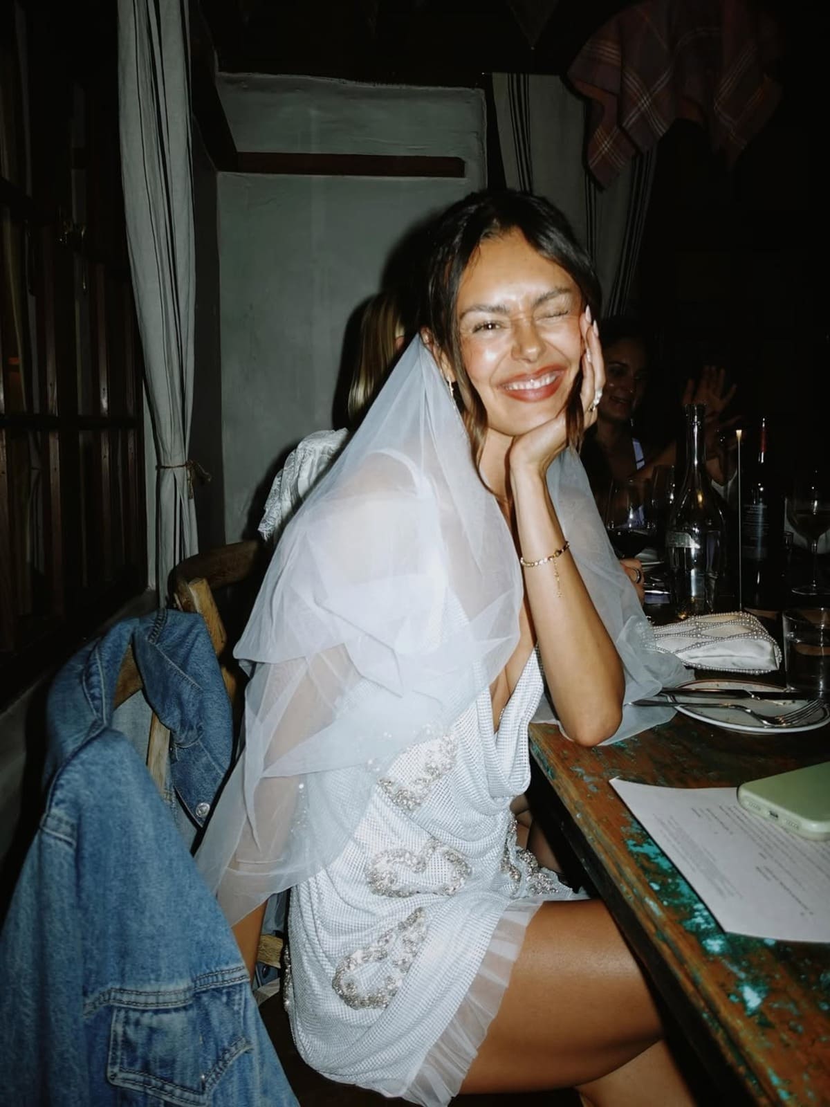 Bride-to-be in a white embellished mini dress and veil, smiling at the dinner table during a stylish Bath hen weekend, one of the best Bath hen do activities for a celebratory evening