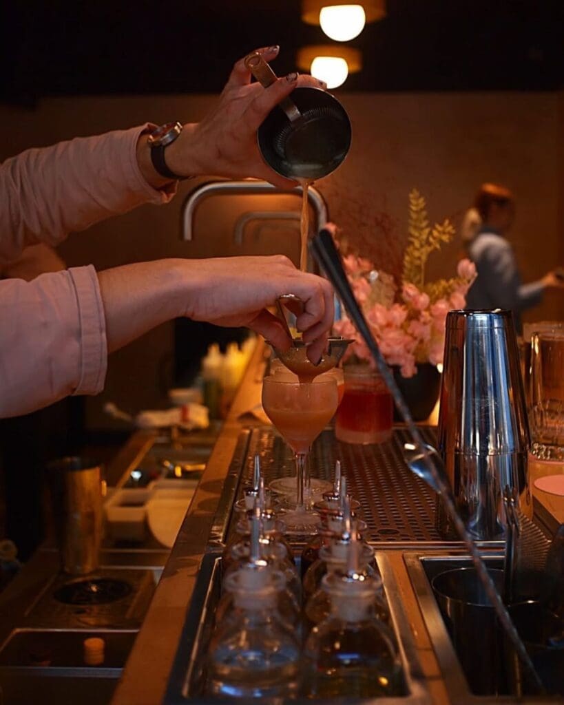Bartender pouring a cocktail at Posie in Manchester, with low lighting, pink flowers and a softly lit bar setup for a stylish Manchester girls’ night.