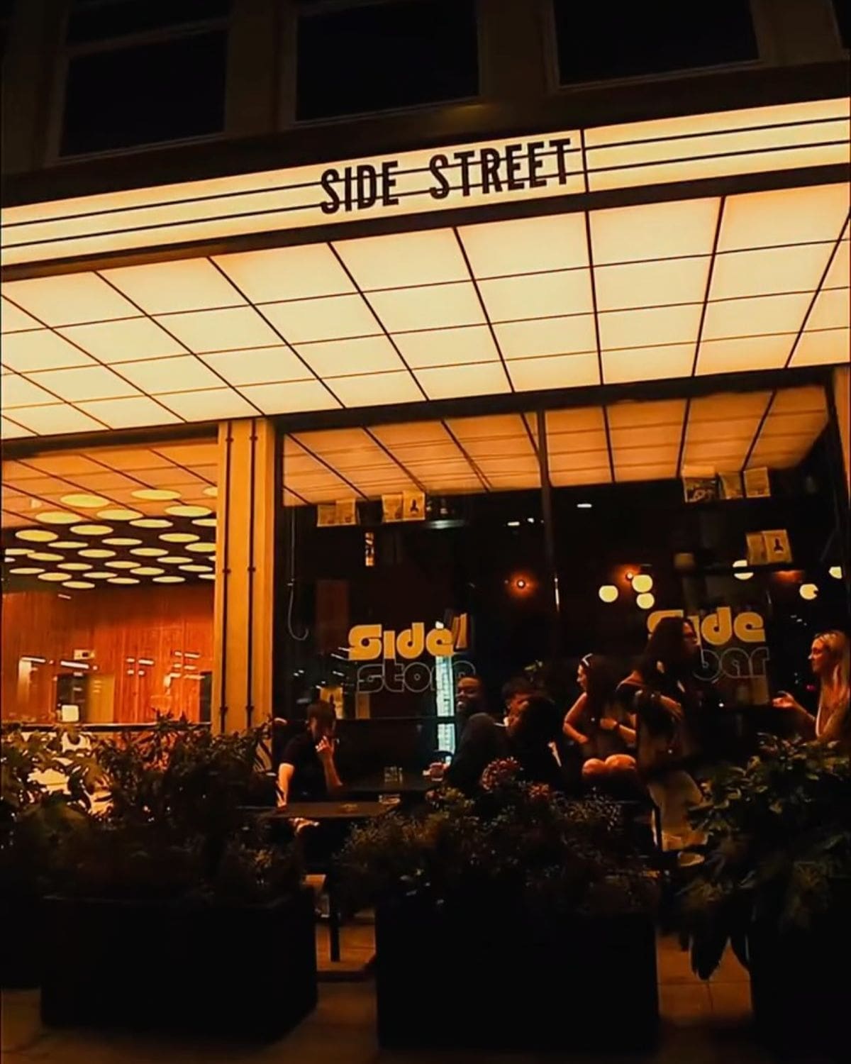 Glowing Side Street sign outside the Manchester bar at night, with warm lighting, indoor-outdoor seating and a buzzy crowd for a girls’ night out in Manchester.