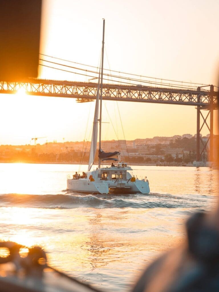 Catamaran sailing on the Tagus River at sunset under Lisbon’s 25 de Abril Bridge, featured as inspiration for Lisbon hen activities and boat hire.