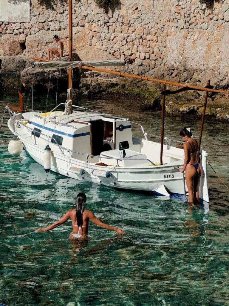 Palma hen activities image of a small white boat moored in clear turquoise water, with two women swimming nearby and rocky coastline in the background.