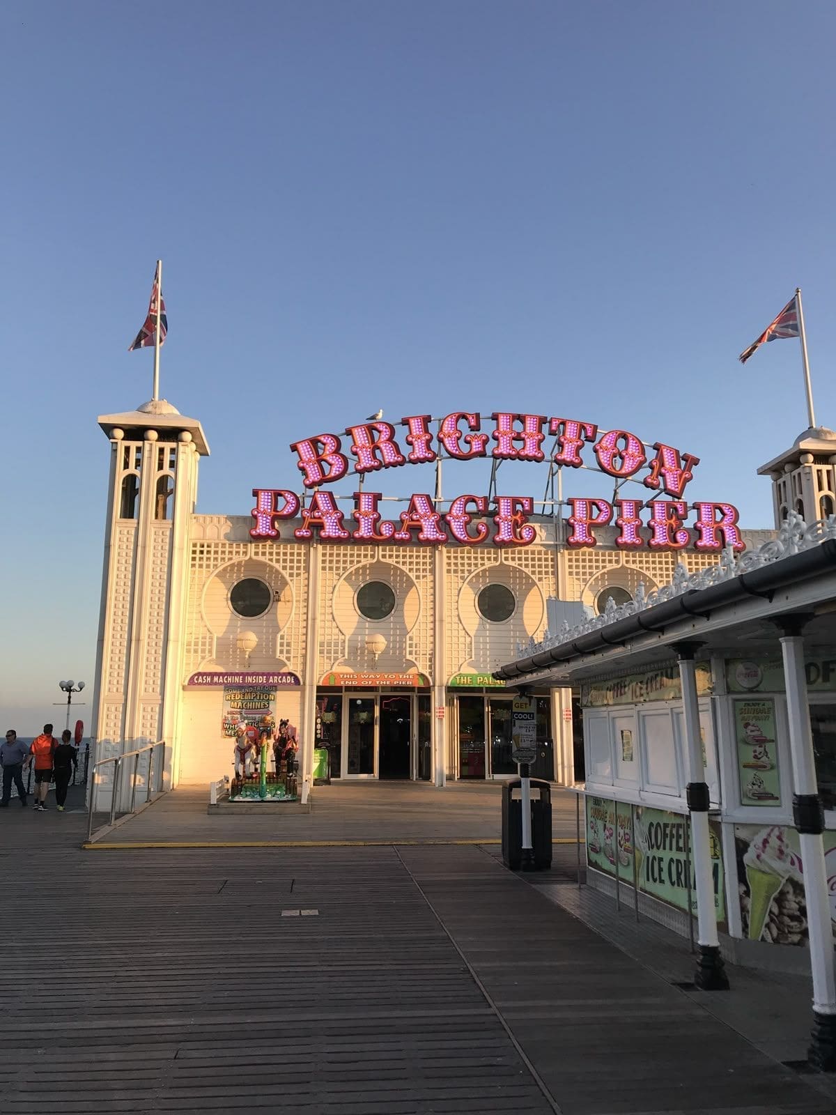 Brighton hen do activities image of the Brighton Palace Pier entrance at sunset, with the sea, wooden boardwalk and retro pier signage in golden evening light.