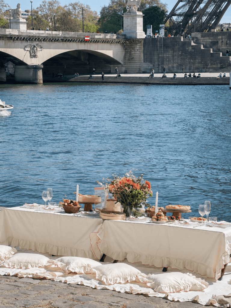 Paris hen do activities image of a styled picnic by the River Seine, with cushions, flowers, food and the Eiffel Tower visible in the background.