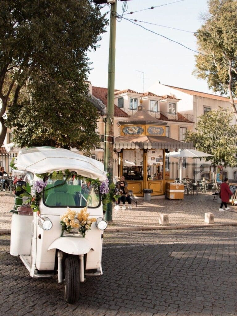 Lisbon hen activities image of a white tuk tuk on a cobbled street, with a kiosk café and sunny neighbourhood square in the background.