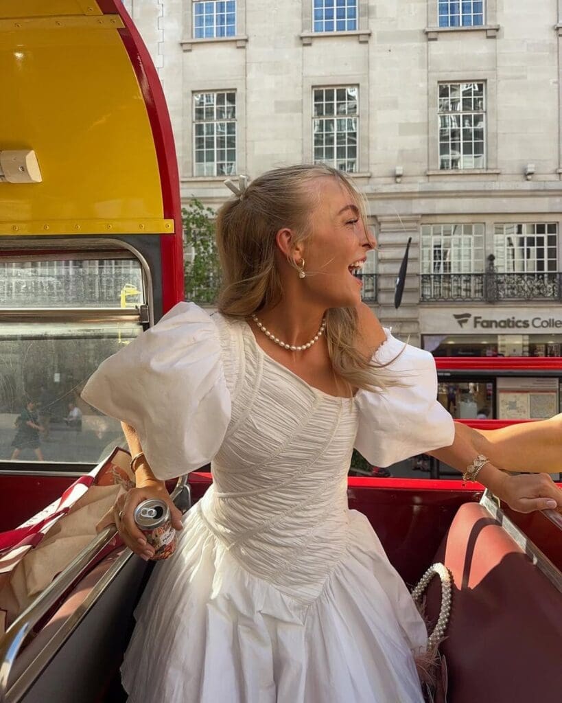 London hen activities inspiration image of a bride-to-be in a white dress sitting on the top deck of an open-top red bus, laughing with London streets behind her.