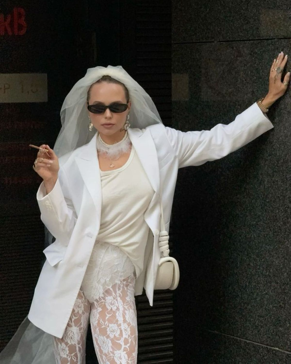London hen activities inspiration image of a bride-to-be in a white blazer, veil, lace trousers and sunglasses, posing against a dark wall in a chic city outfit.