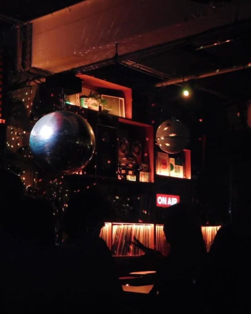 The dark, retro interior of Café 1001 in Shoreditch, with disco balls, glowing red shelves and an illuminated On Air sign — a moody late-night spot for a London night out.