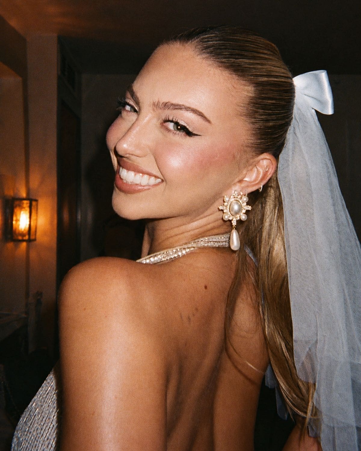 A bride-to-be smiling over her shoulder in a veil, statement pearl earrings and party-ready evening look — a stylish cover image for where to go out-out on a Manchester hen weekend.