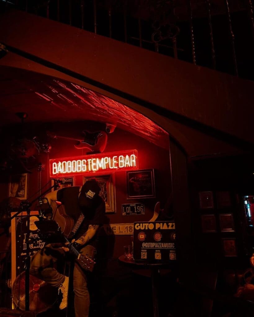 A live musician performing inside Bad Bobs Temple Bar in Dublin, framed by dark interiors and a glowing red neon sign — a late-night bar for live music and going out-out in the city.